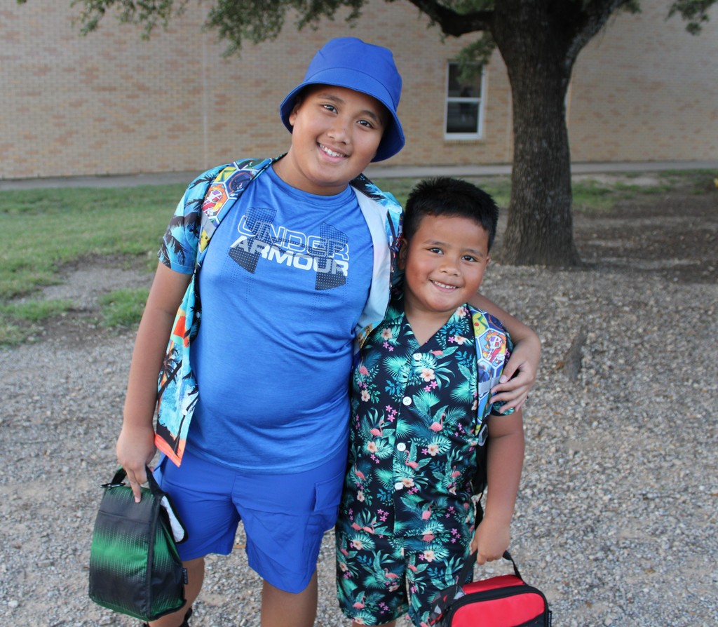 two students in tropical shirts and hat smile outside school