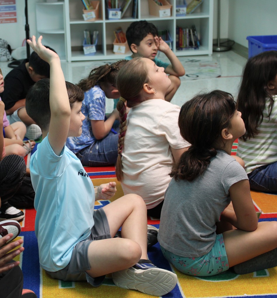 Students sitting on carpet raise hands