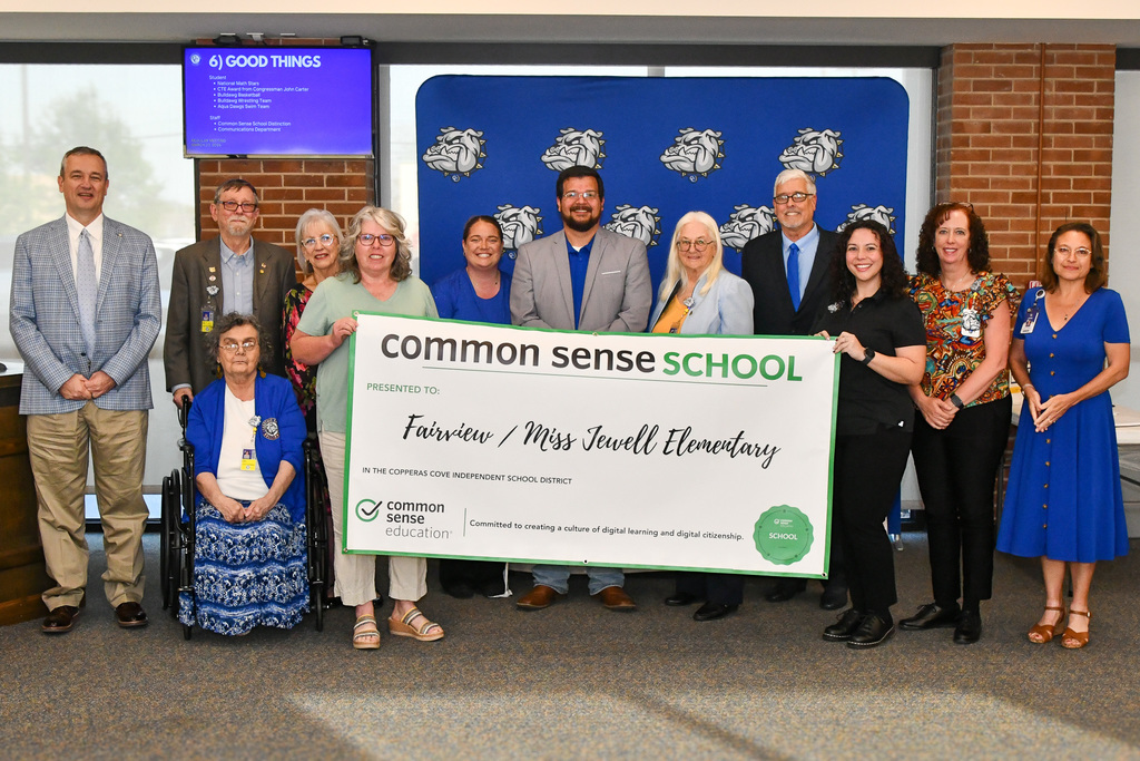 A group of teachers and trustees holding a banner and smiling in front of a backdrop