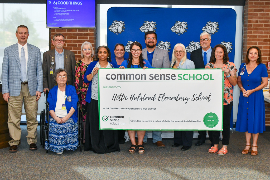 A group of teachers and trustees holding a banner and smiling in front of a backdrop