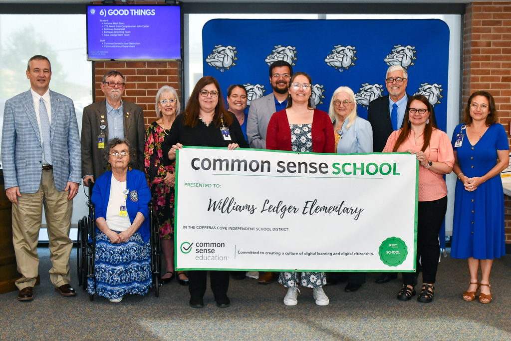 A group of teachers and trustees holding a banner and smiling in front of a backdrop