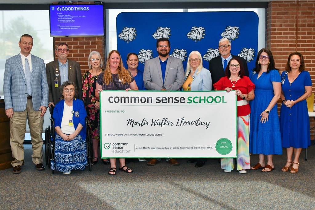 A group of teachers and trustees holding a banner and smiling in front of a backdrop