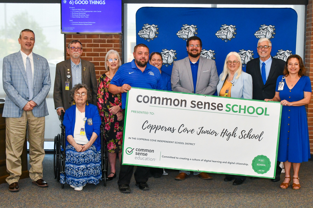 A group of teachers and trustees holding a banner and smiling in front of a backdrop