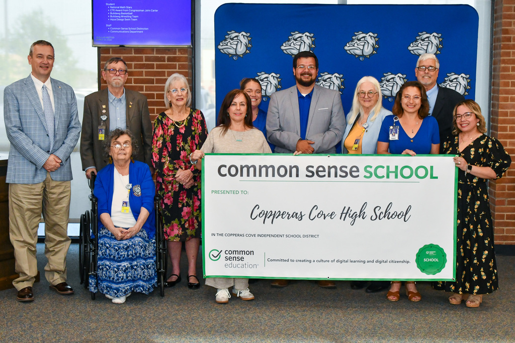 A group of teachers and trustees holding a banner and smiling in front of a backdrop