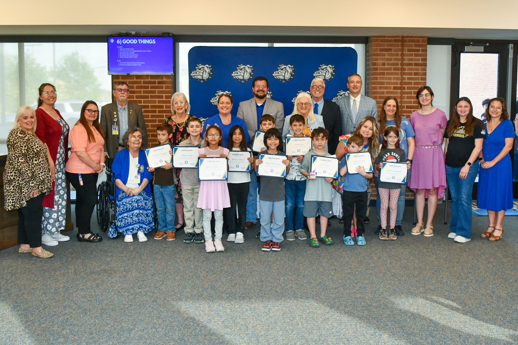 A group of students holding awards and adults smiling in front of a backdrop