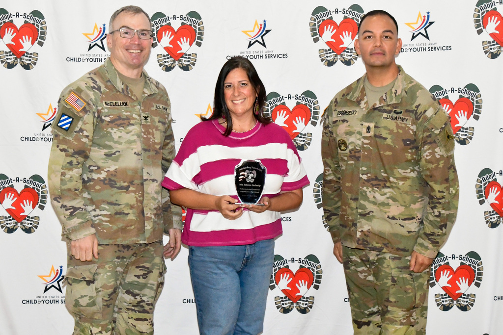 Two soldiers and a woman smiling in front of a back drop holding an award