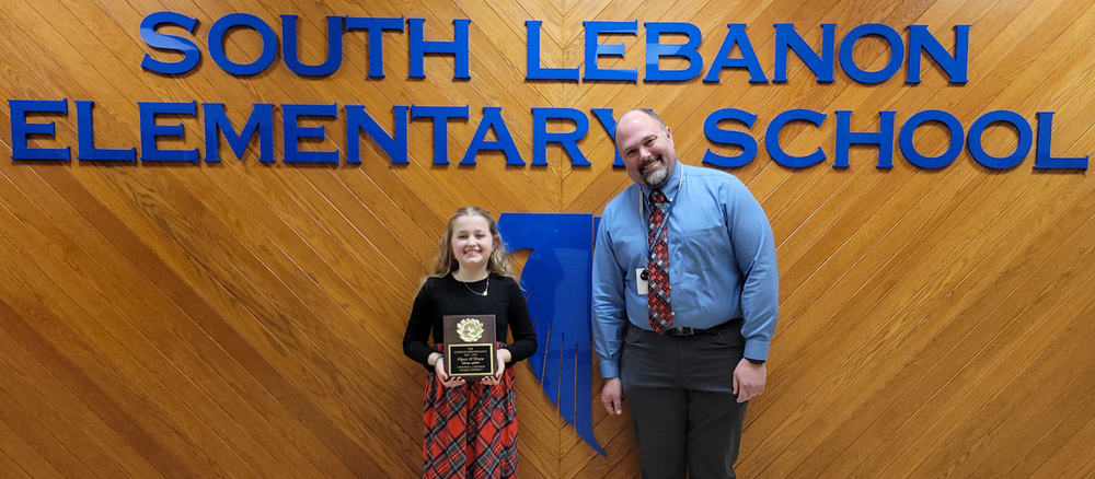 An elementary school student holds a Student of the Year award plaque while standing beside a school administrator in front of the South Lebanon Elementary School wall sign.