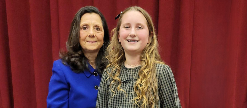 An elementary school student smiles while standing beside the Board president. They are posing in front of red stage curtains.