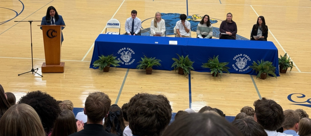 A speaker stands at a podium in a high school gymnasium while six people sit behind a long table draped in blue Cedar Crest High School cloths. An audience is seated in the foreground.