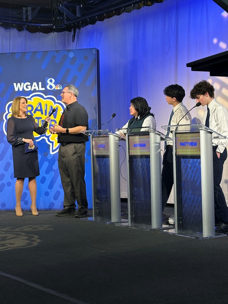 Female reporter interviewing male advisor in front of WGAL 8 Brainbusters sign with three male teens nreaby behind podiums