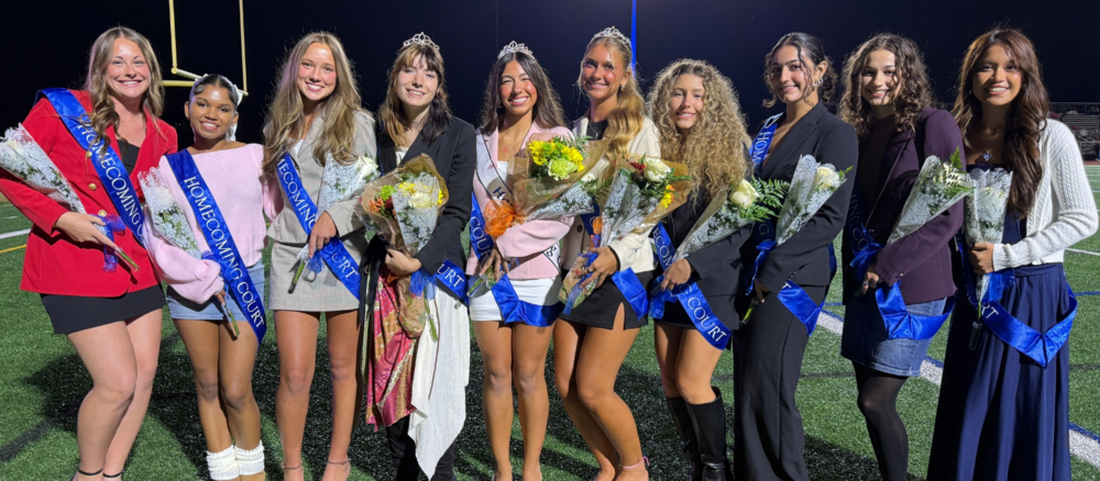 Ten students wearing blue homecoming court sashes pose together on a football field at night, several holding bouquets of flowers and four wearing tiaras.