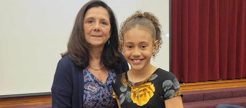 An elementary school student smiles while standing beside a woman during a school recognition event. They are indoors in front of a projection screen and stage curtains.