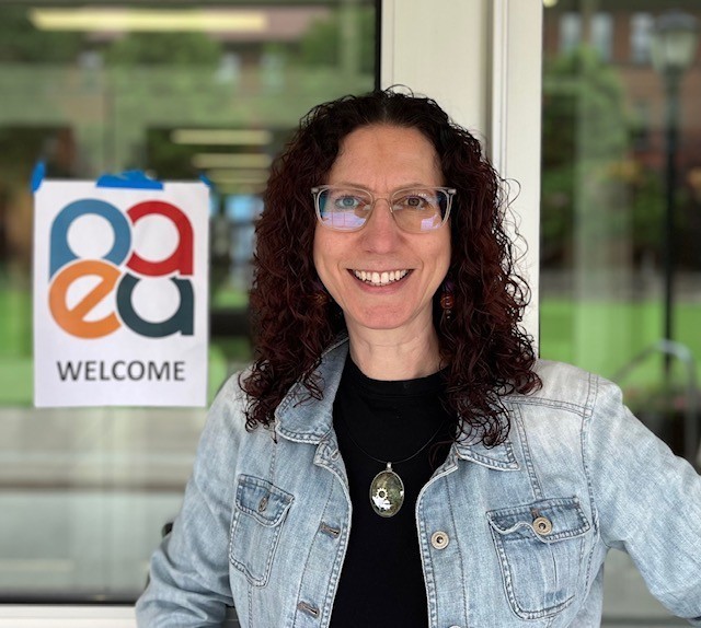 Andrea Klein standing in a jean jacket in front of glass doors with a sign "PEAA Welcome"