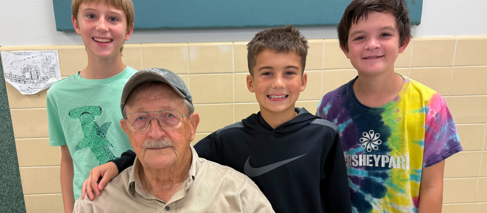 Three elementary-aged students pose with an older adult indoors near a beige-tiled wall; one student has an arm around the seated adult’s shoulder.