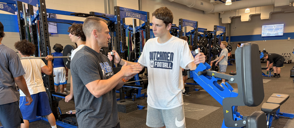 A Cedar Crest High School strength and conditioning coach talks with a student-athlete beside weight-training equipment in the school’s renovated weight room, where other student-athletes are exercising in the background.