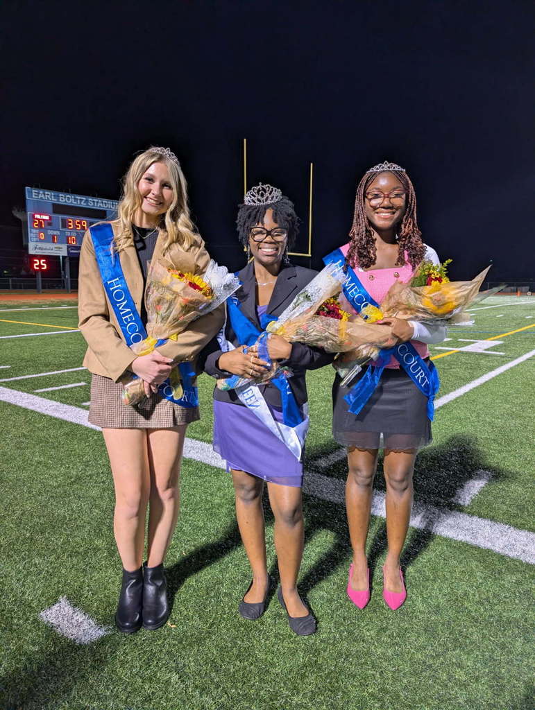 Three girls in short dresses on the football field, wearing sashes and crowns and carrying flowers