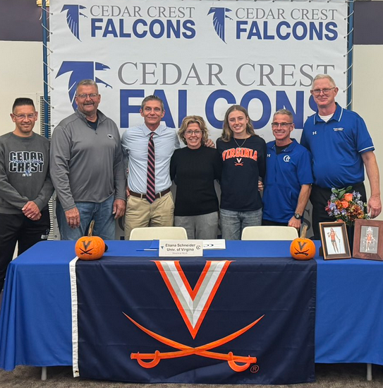 Five male coaches and one female coach with Ellie Schneider stand in front of Cedar Crest Falcons sign and Univ. of Virgina banner.