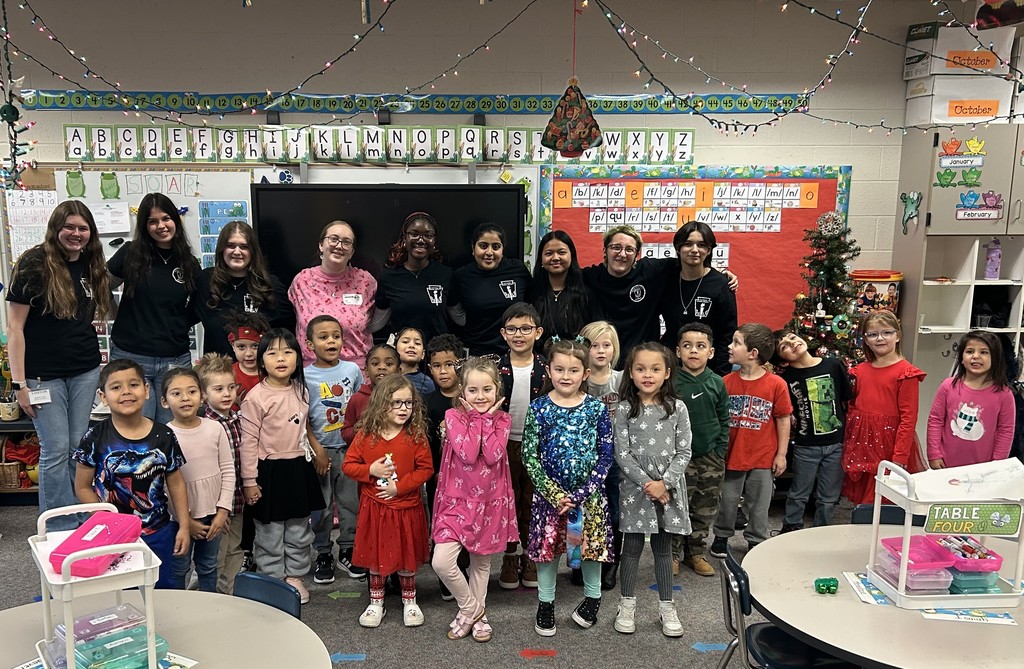 Nine high school students stand in a line behind 19 Kindergarten students in their classroom