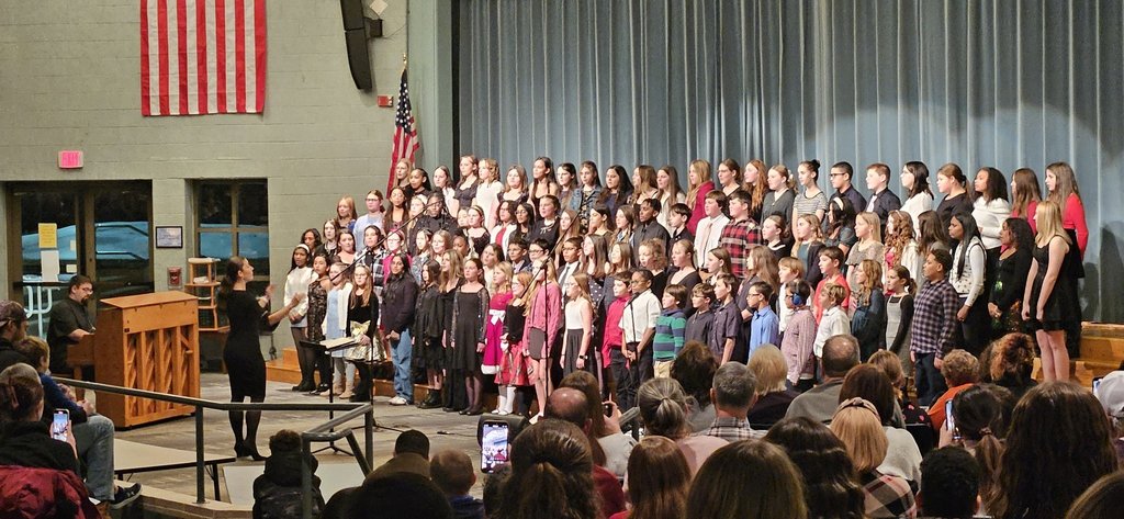A teacher directing about 30 6th grade students singing at a holiday concert with part of the audience in the foreground