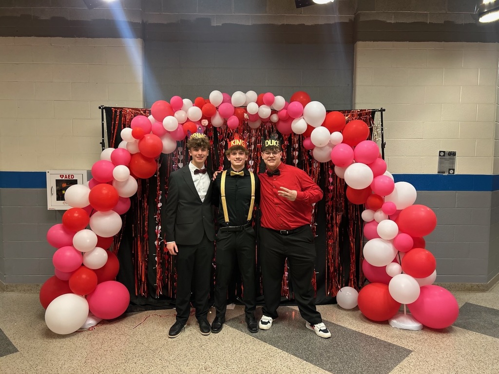 three high school senior boys in suits wearing crowns while standing under and archway of red balloons