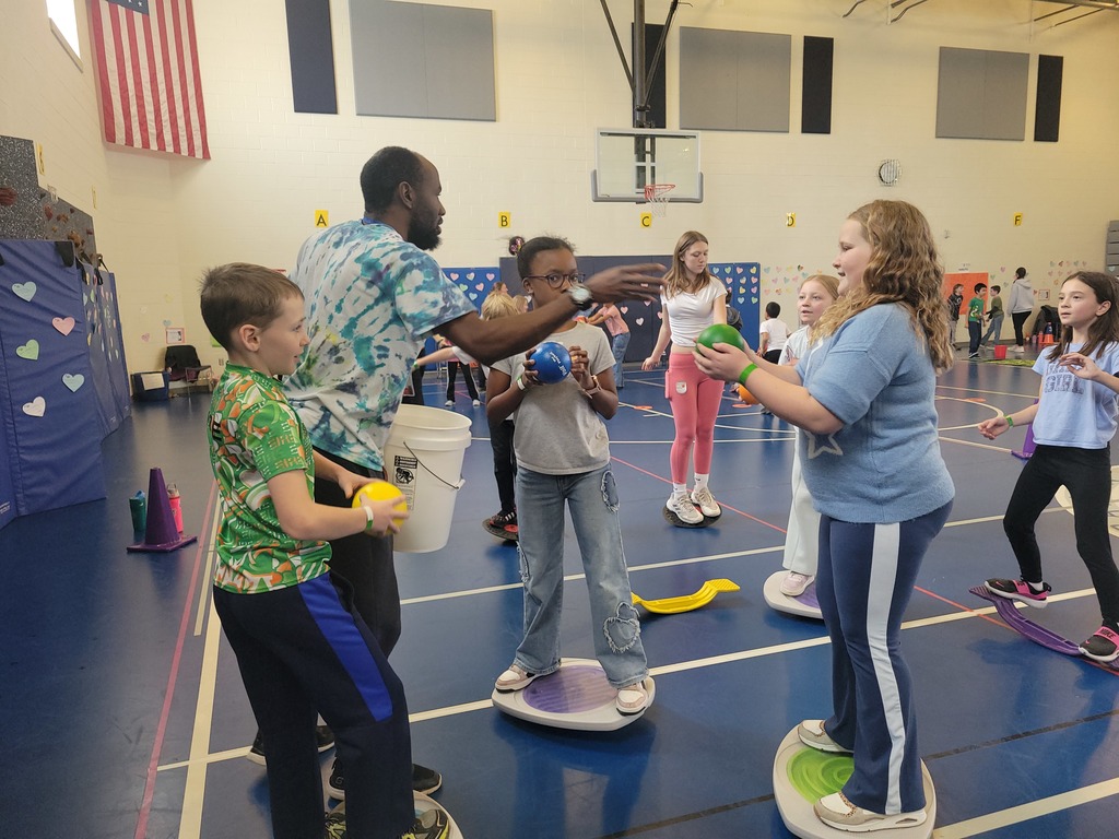 Adult volunteer hands out balls for elementary kids to toss in a bucket while the balance on floor teeter-totters
