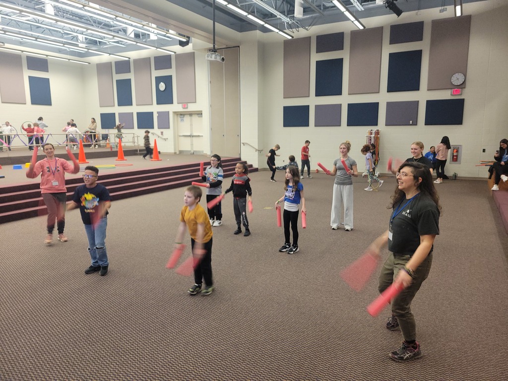 group of children and adults in a large empty room holding flexible foam sticks and following an exercise dance routine