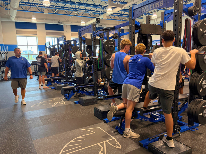Cedar Crest High School football student-athletes train in the renovated weight room, using squat racks and strength-training equipment while a coach walks through the facility.
