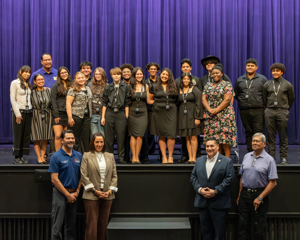 Students, teachers and superintendent stand on stage and school board trustees stand in front of stage at Roy Miller High School Ribbon Cutting event.