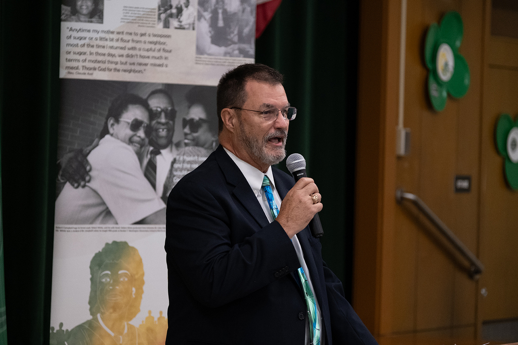 Marty Bell, a school board member, speaks into a handheld microphone while standing indoors at a school celebration event. He wears a dark suit jacket, white shirt, and colorful tie.
