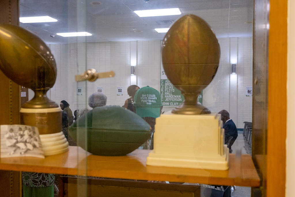 A glass display case shows vintage football trophies and memorabilia from Solomon M. Coles High School, including a plaque and a green football.