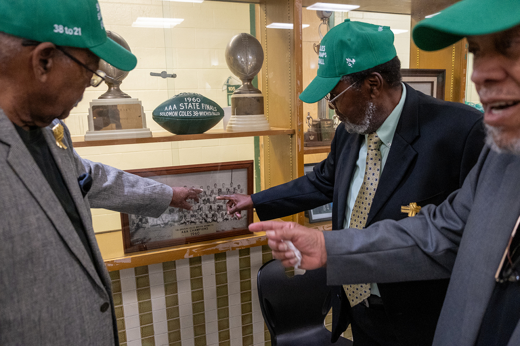 Three alumni wearing green Solomon M. Coles caps point to individuals in a framed 1960 championship football team photo displayed in a trophy case. Above the photo sit vintage trophies and a green football labeled “1960 AAA State Finals – Solomon Coles 38, Wichita Falls 21.”