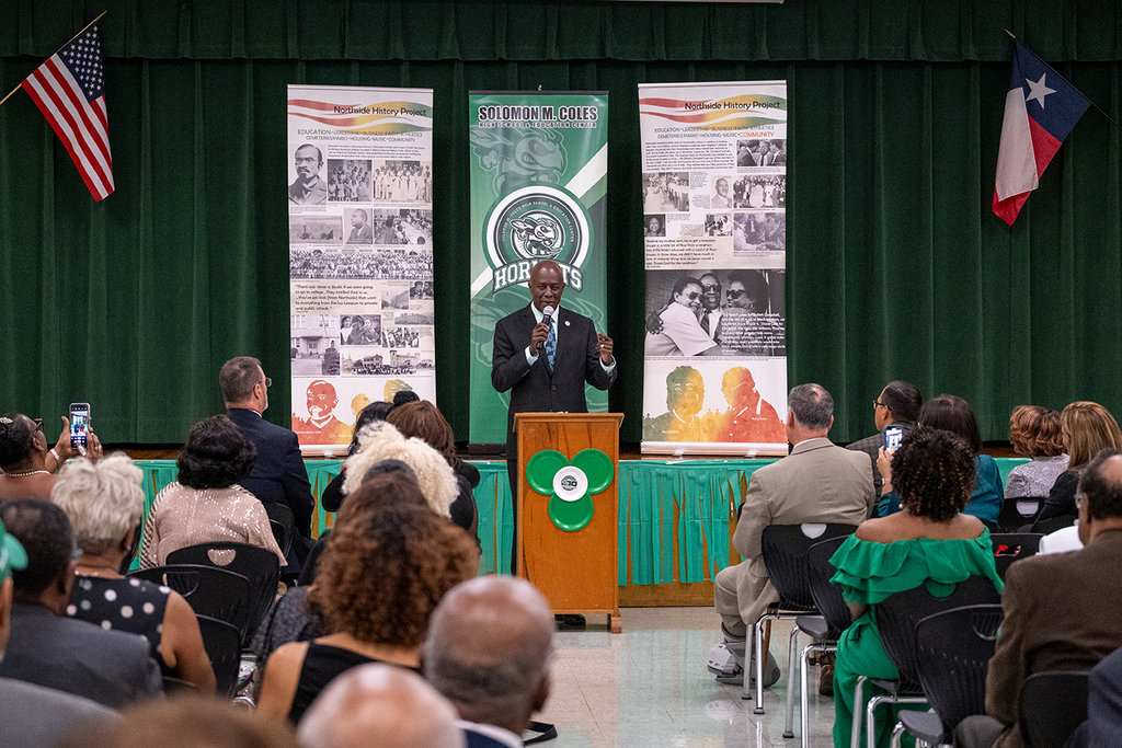 Dr. Bruce Wilson speaks at a podium during the Solomon M. Coles 100th anniversary celebration. He wears a dark suit and tie, holding a microphone as he addresses the audience seated before him.