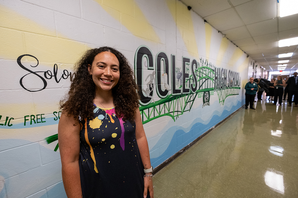 A woman smiles while standing in front of a newly unveiled mural in a school hallway during the Solomon M. Coles 100th anniversary celebration.