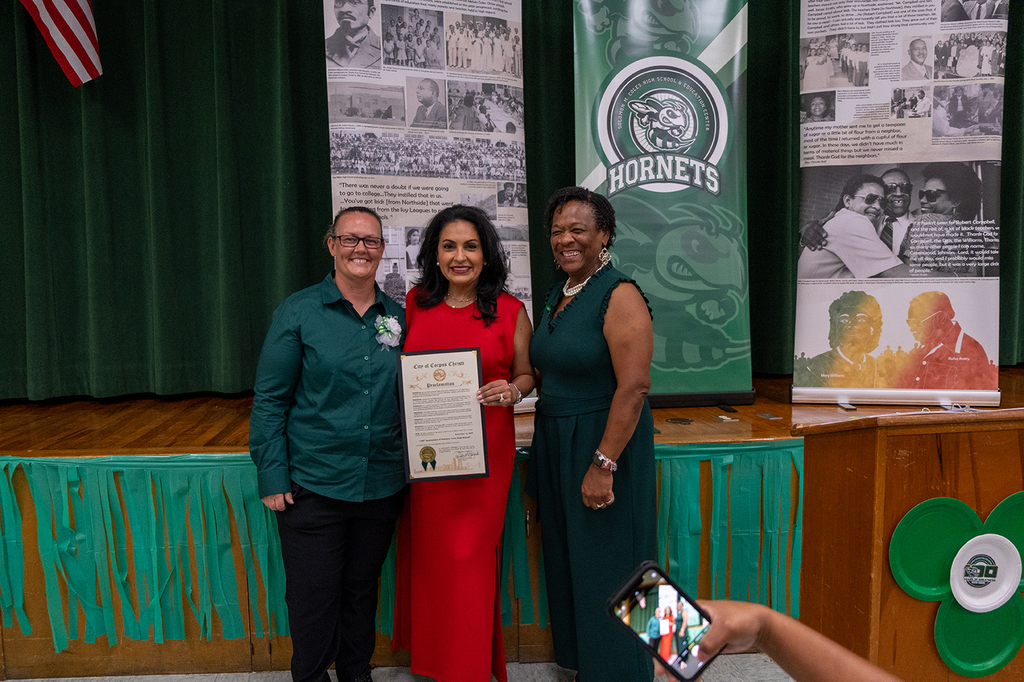 Three women pose together indoors at a school celebration event. The woman in the center, wearing a bright red dress, holds a framed proclamation from the City of Corpus Christi. The other two women, dressed in dark green attire, stand on either side of her smiling.