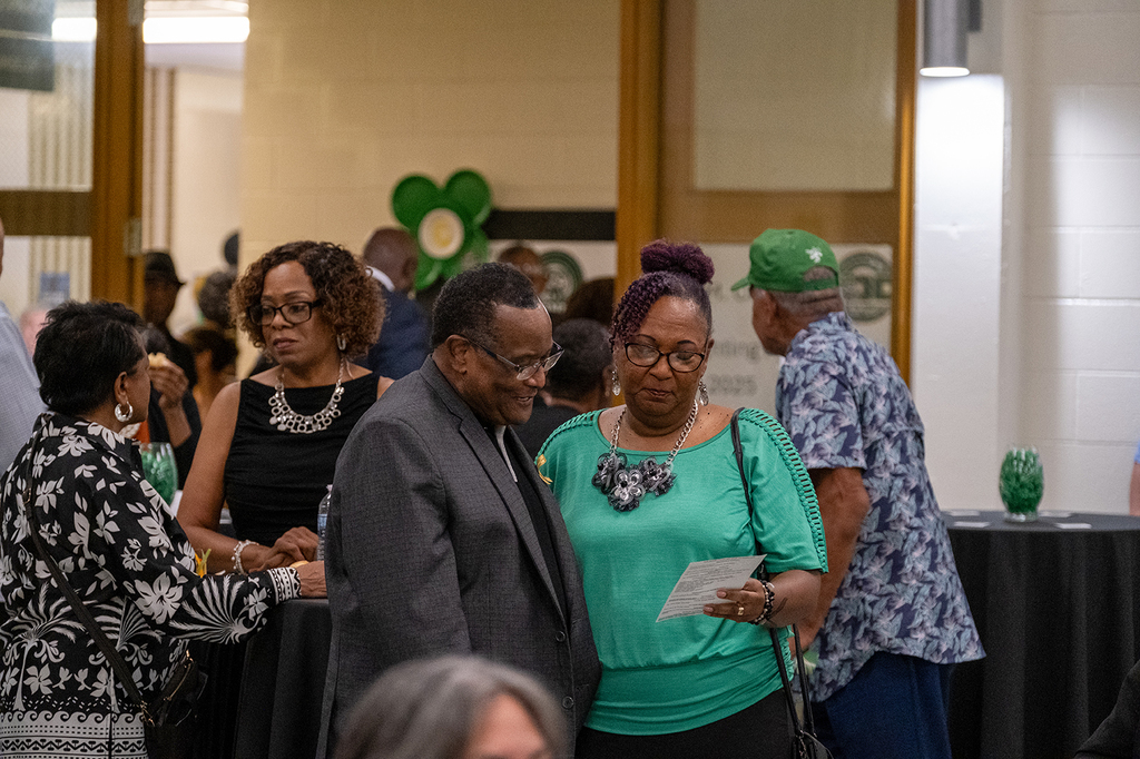 Attendees mingle and chat during the Solomon M. Coles 100th anniversary celebration. A man in a dark suit and a woman in a green blouse read a program together in the foreground