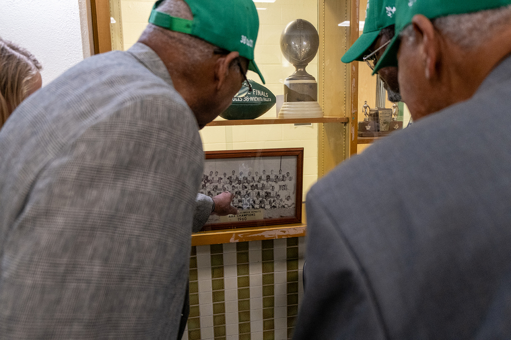 Two alumni wearing green Solomon M. Coles caps lean in to look at a framed black-and-white team photo from 1960 displayed in a trophy case.
