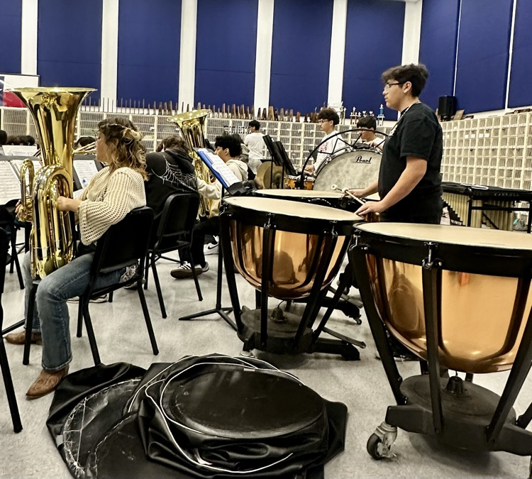 Band students practicing for a concert; percussion and tuba sections are focused while a student plays timpani drums in the foreground.