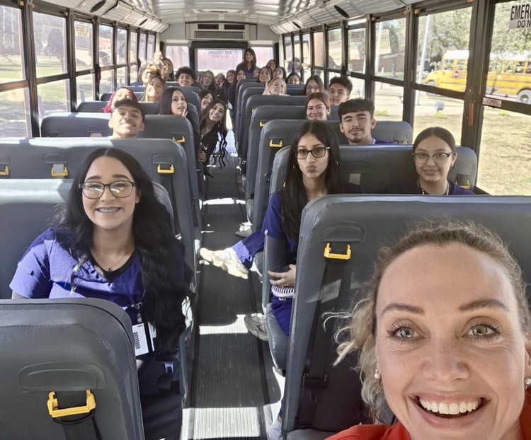 Smiling teacher takes a selfie on a school bus filled with CTE Phlebotomy students wearing navy scrubs, all seated and ready to head out for their ID badge trip.
