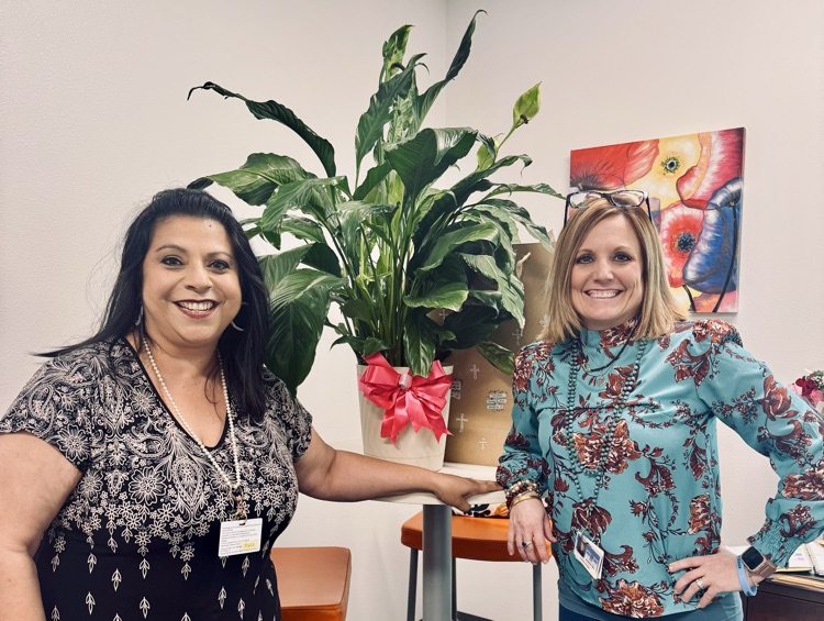 Dr. Melissa Hernandez and Ms. Filla smiling together in an office space, standing beside a large green plant wrapped with a pink bow, with colorful artwork hanging on the wall behind them.