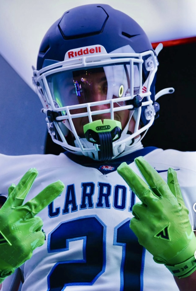A Carroll High School football player wearing a white jersey with blue lettering and a navy helmet poses confidently with both hands raised, flashing peace signs in bright neon green gloves.