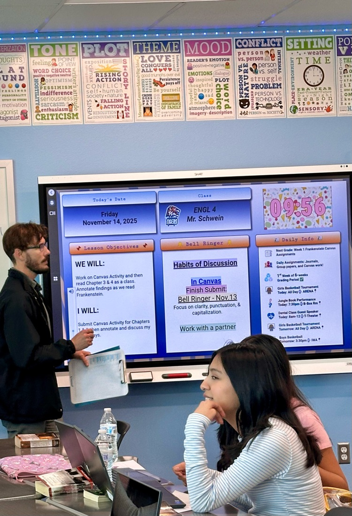 Alt Text: Students in English class read and annotate Frankenstein at their desks and, some using laptops, with posters, a smartboard lesson screen, and classroom decorations visible in the background.