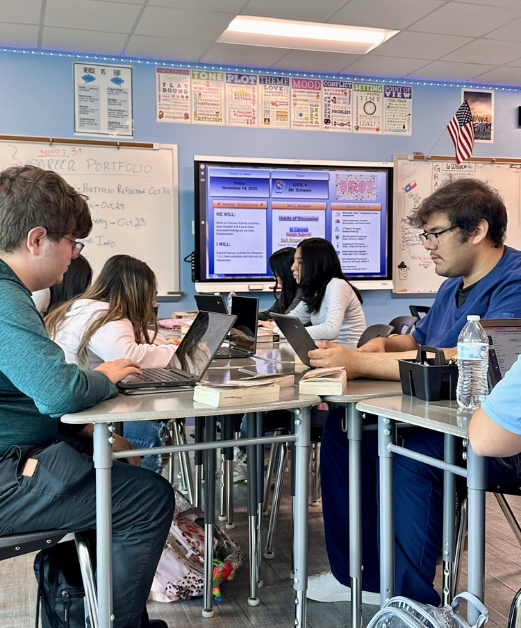 Alt Text: Students in English class read and annotate Frankenstein at their desks and, some using laptops, with posters, a smartboard lesson screen, and classroom decorations visible in the background.