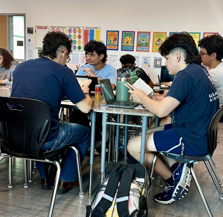 Alt Text: Students in English class read and annotate Frankenstein at their desks and, some using laptops, with posters, a smartboard lesson screen, and classroom decorations visible in the background.