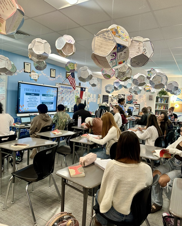 Alt Text: Students in English class read and annotate Frankenstein at their desks and, some using laptops, with posters, a smartboard lesson screen, and classroom decorations visible in the background.