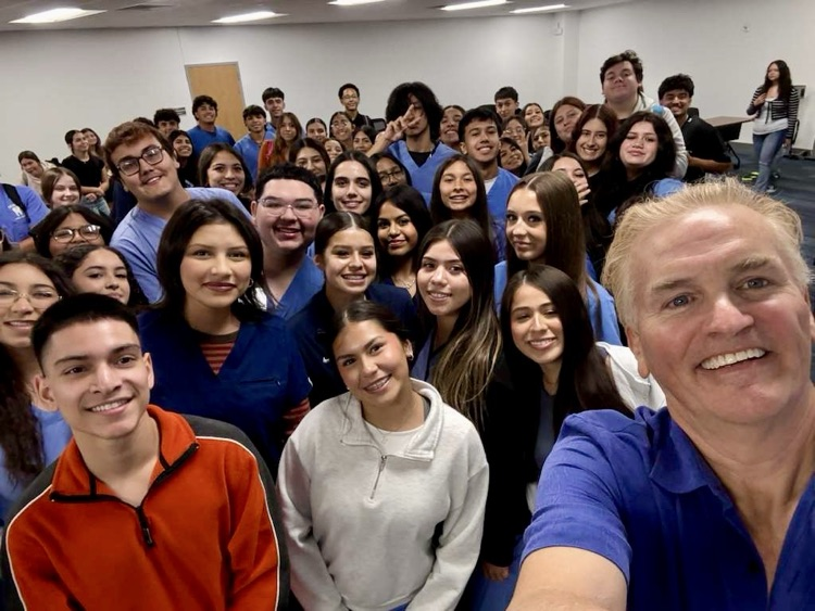 A dentist stands at the front of a classroom speaking to dental students seated in scrubs, watching a presentation on a screen.