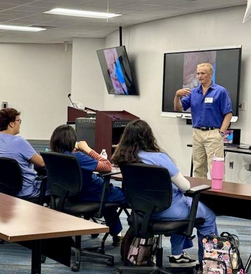 A dentist stands at the front of a classroom speaking to dental students seated in scrubs, watching a presentation on a screen.