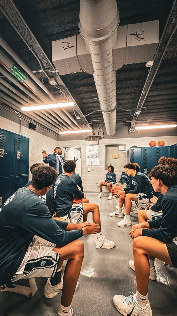 Short Alt Text: Basketball team in the locker room listening to coach after a big win.