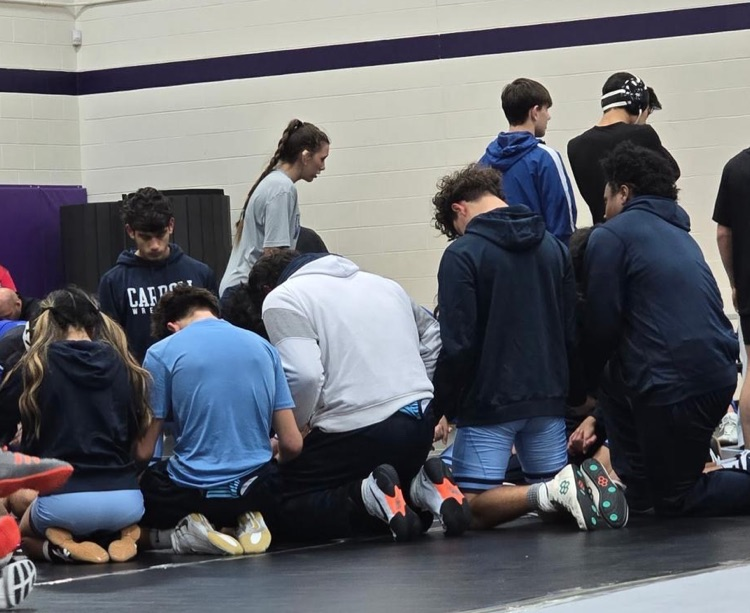 Alt Text: Carroll High School wrestlers are kneeling together in a tight huddle on the gym floor, preparing before their matches.