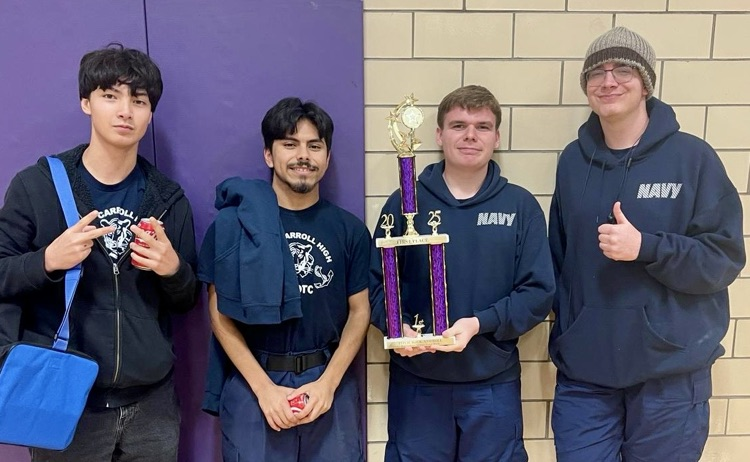 ⸻ Alt Text: Image 1: Four NJROTC cadets stand together indoors wearing Carroll and Navy hoodies. They’re smiling and holding a tall purple-and-gold trophy with a “1st place” plaque.