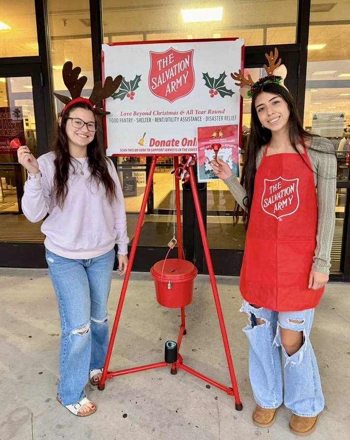 ⸻ Alt Text: Two Junior Council students wearing reindeer antlers ring bells beside a Salvation Army red kettle outside JCPenney.
